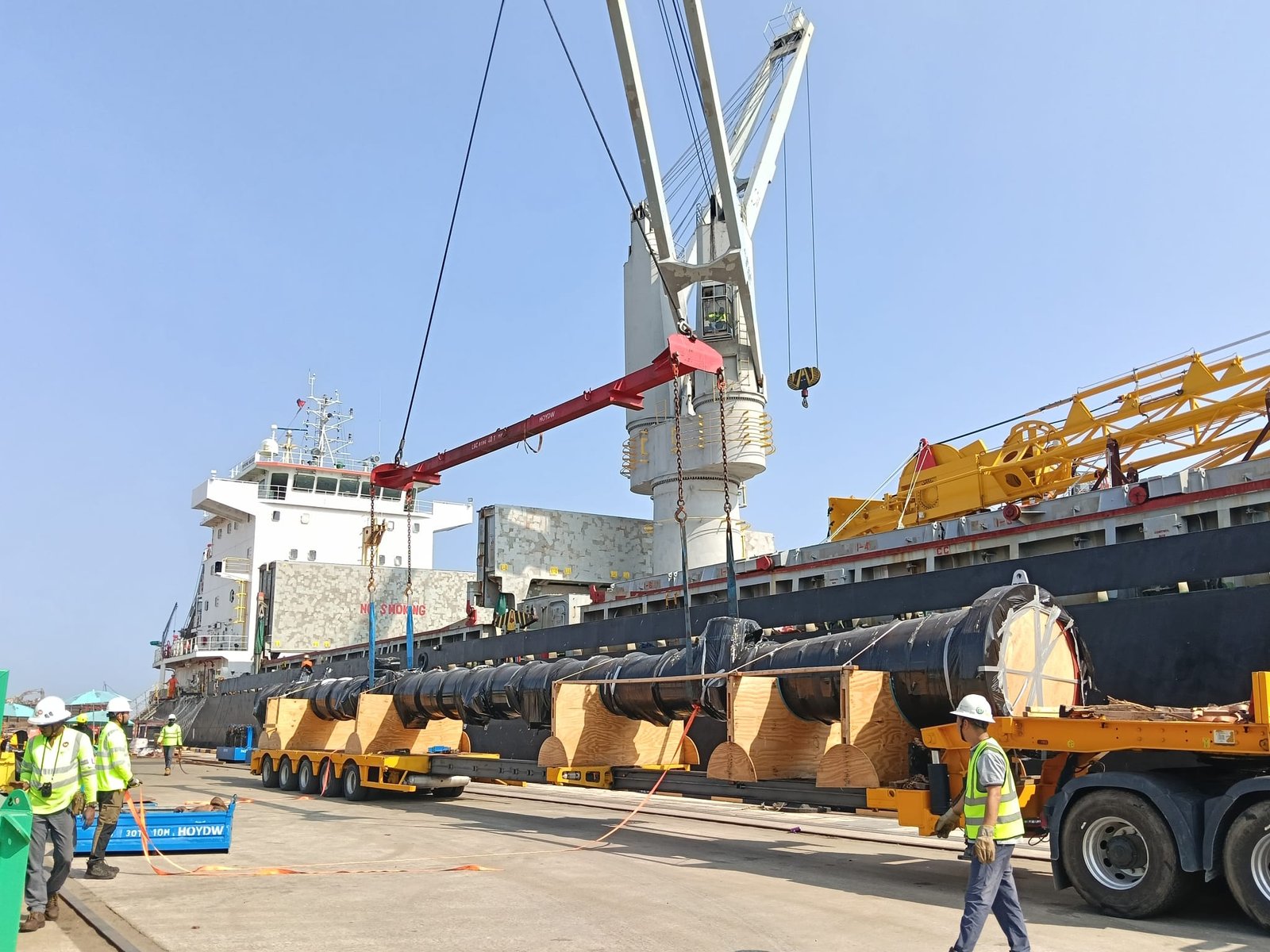 Ship crane lowering large pipe sections onto an SPMT trailer at a project cargo berth
